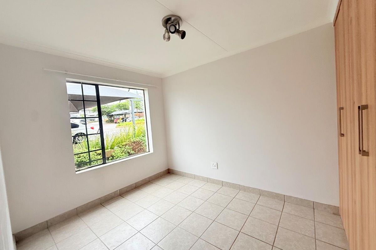 A bright, empty bedroom featuring neutral floor tiles, built-in wooden cupboards, and a window with security bars overlooking the complex exterior.