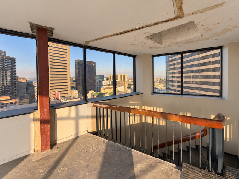 An empty penthouse living area featuring floor-to-ceiling windows with city views, a central staircase, and significant ceiling finish deterioration requiring renovation.