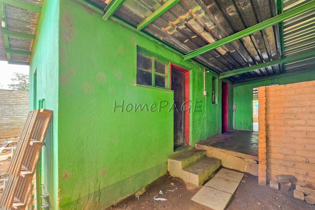 Exterior view of a basic dwelling featuring a covered walkway, concrete steps, and painted masonry walls with a corrugated metal roof structure.