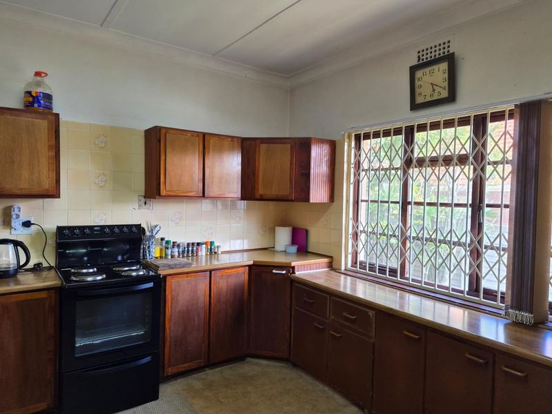 A dated kitchen featuring wooden cabinetry, laminate countertops, and a tiled backsplash, equipped with a freestanding stove and a window with security bars.