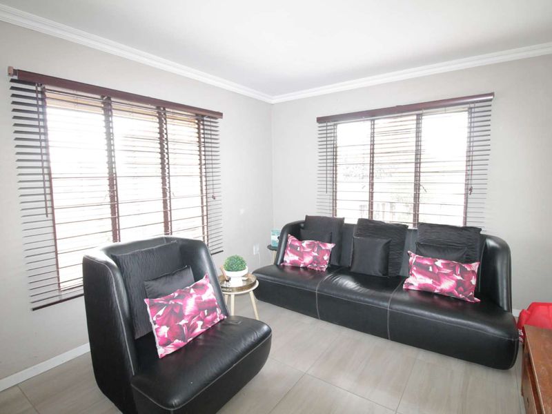 A bright living room featuring neutral wall tones, tiled flooring, and dual windows fitted with dark venetian blinds.