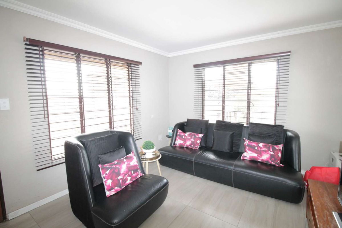 A bright living room featuring neutral wall tones, tiled flooring, and dual windows fitted with dark venetian blinds.