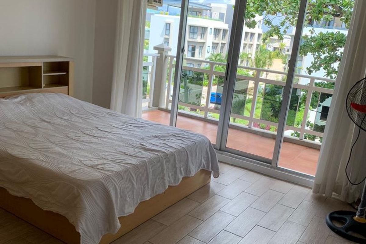 A bright bedroom featuring wood-look tile flooring, a wall-mounted air conditioning unit, and large sliding glass doors opening onto a private balcony.