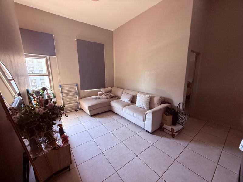 A living room featuring neutral wall tones, tiled flooring, and large windows fitted with roller blinds, offering a functional and bright living space.