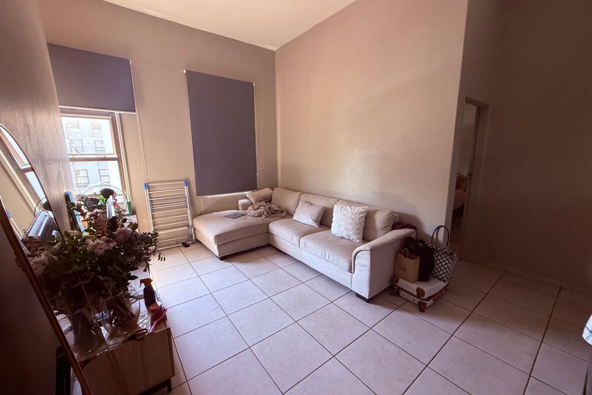 A living room featuring neutral wall tones, tiled flooring, and large windows fitted with roller blinds, offering a functional and bright living space.