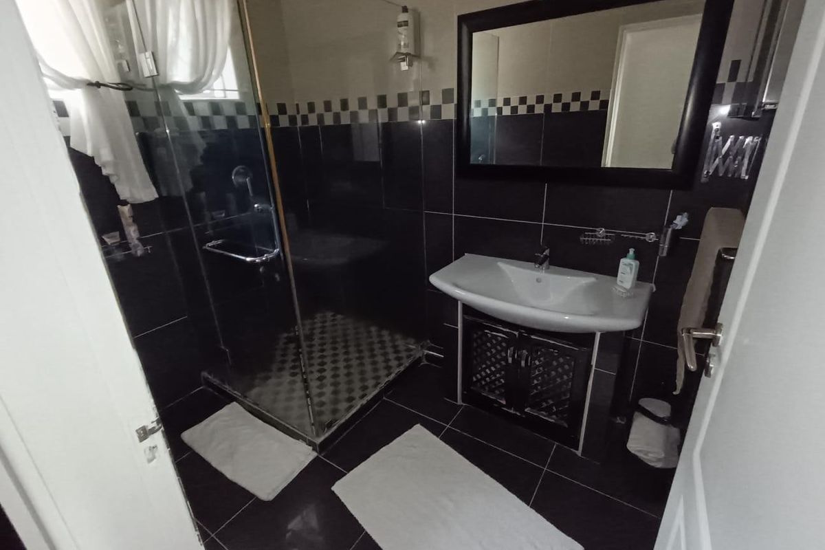 A compact bathroom featuring dark tiled walls and flooring, a glass shower enclosure, and a wall-mounted vanity basin with a framed mirror.