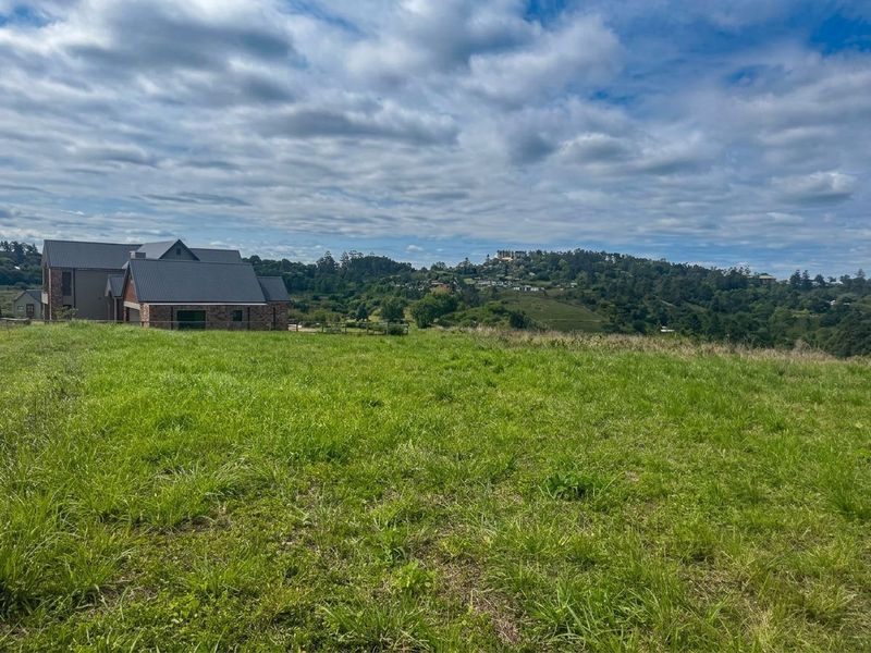 A scenic view of a vacant residential plot featuring rolling green hills and an adjacent modern home under a cloudy sky.