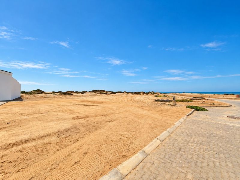 A vacant residential plot featuring sandy terrain, adjacent to a paved street with coastal views in the distance.