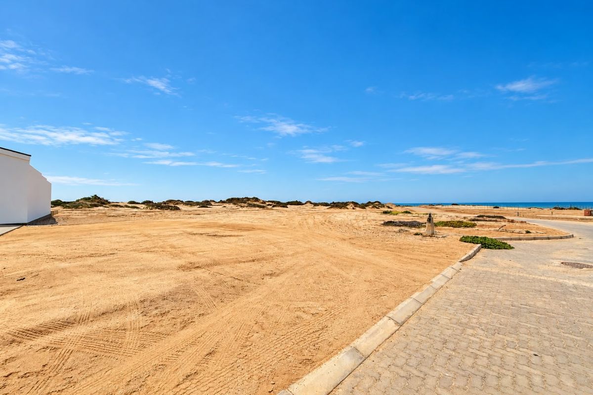 A vacant residential plot featuring sandy terrain, adjacent to a paved street with coastal views in the distance.