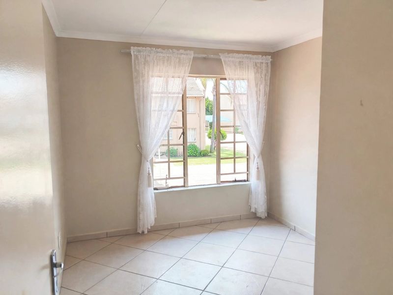 A bright, empty bedroom featuring neutral wall tones, tiled flooring, and a large metal-framed window overlooking the complex grounds.