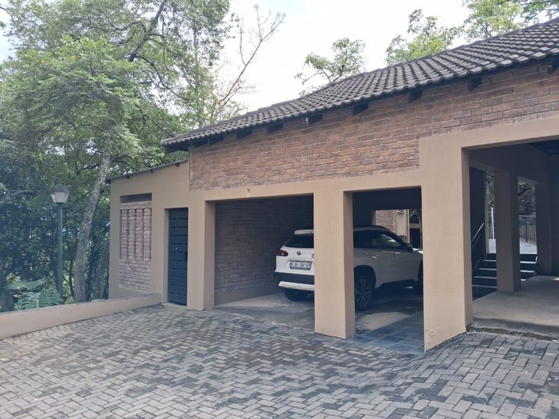 A well-maintained apartment complex exterior featuring a brick facade, tiled roof, and covered parking bays with paved driveway access.