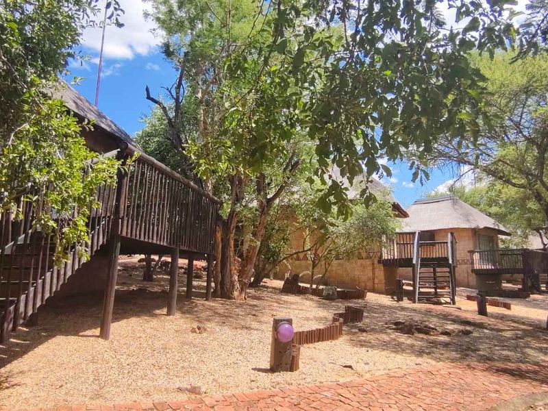 Exterior view of rustic lodge-style units featuring thatched roofs, raised timber decks, and natural stone cladding set within a wooded garden landscape.