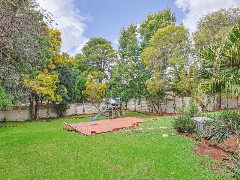 A spacious backyard featuring a large lawn, mature trees, a brick-paved patio area, and a precast concrete boundary wall.