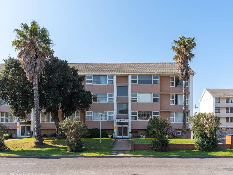 Front elevation of a multi-story apartment building featuring a brick facade, tiled roof, and established garden with palm trees.