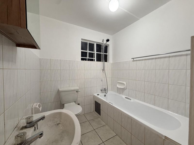 A functional bathroom featuring a bathtub with shower mixer, pedestal basin, and toilet, finished with neutral wall and floor tiling.