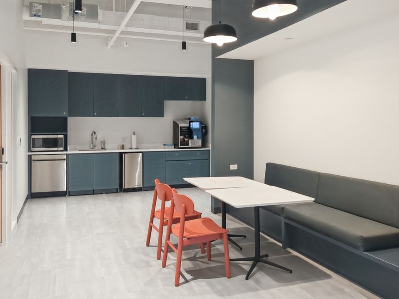 Modern office kitchenette featuring dark blue cabinetry, stainless steel appliances, integrated sink, and a built-in bench seating area with pendant lighting.