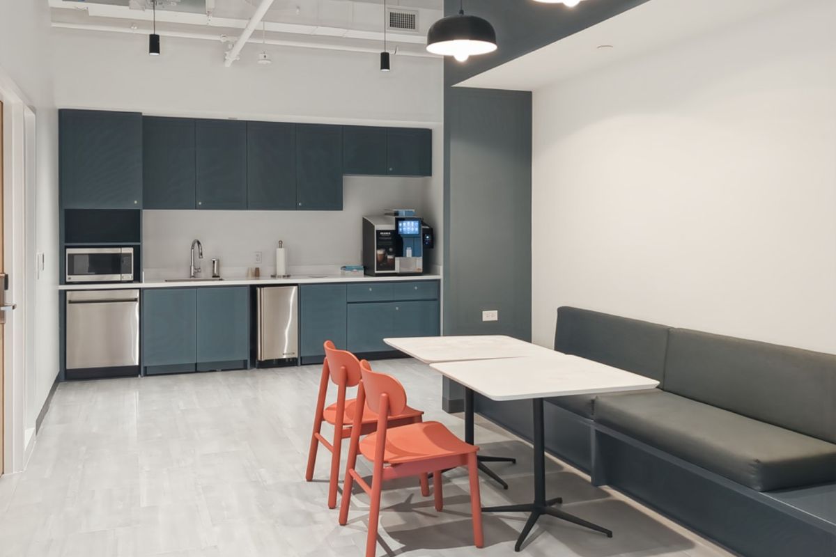 Modern office kitchenette featuring dark blue cabinetry, stainless steel appliances, integrated sink, and a built-in bench seating area with pendant lighting.