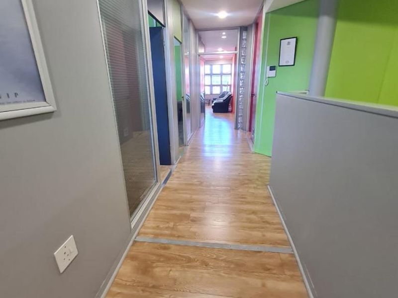 A bright office hallway featuring laminate wood flooring, glass partition walls, and recessed ceiling lighting, providing a clean and professional circulation space.