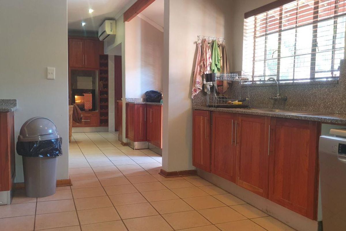 A functional kitchen featuring wooden cabinetry, granite countertops, tiled flooring, and a double sink, with an adjacent scullery area visible in the background.