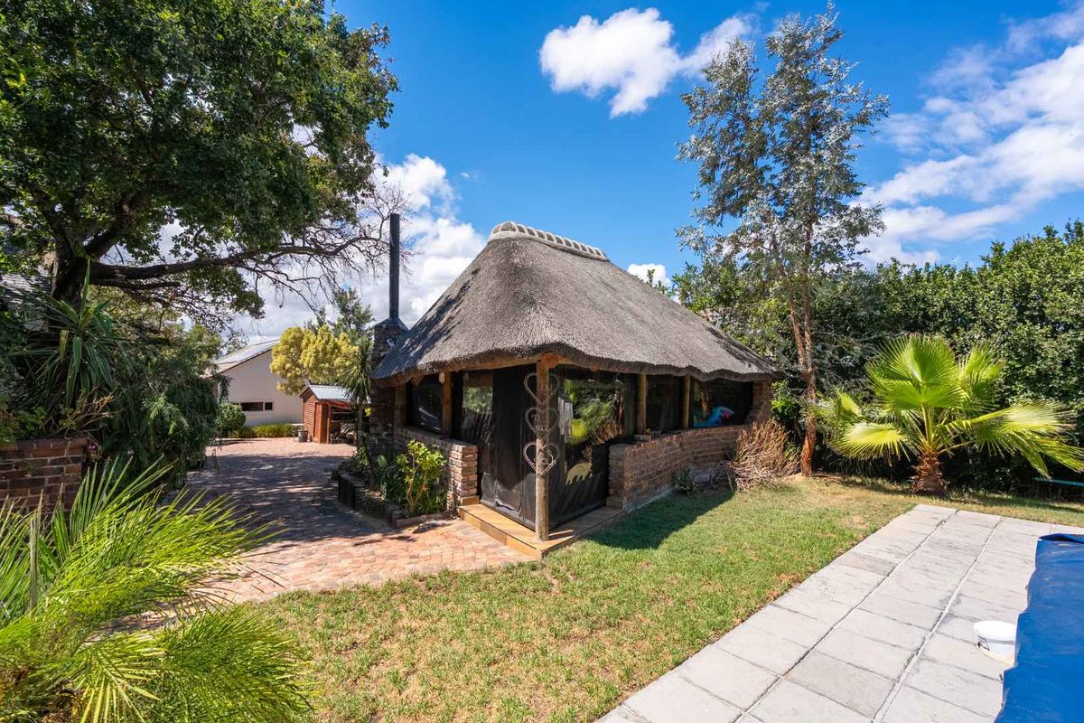 A detached thatched-roof lapa or outdoor entertainment area situated in a landscaped garden, featuring brick construction and an adjacent paved patio area.