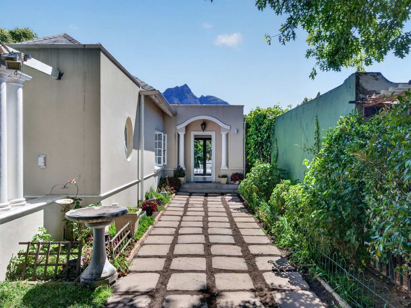 A well-maintained rear exterior view featuring a paved walkway leading to a columned entrance, bordered by a garden wall and mature greenery.