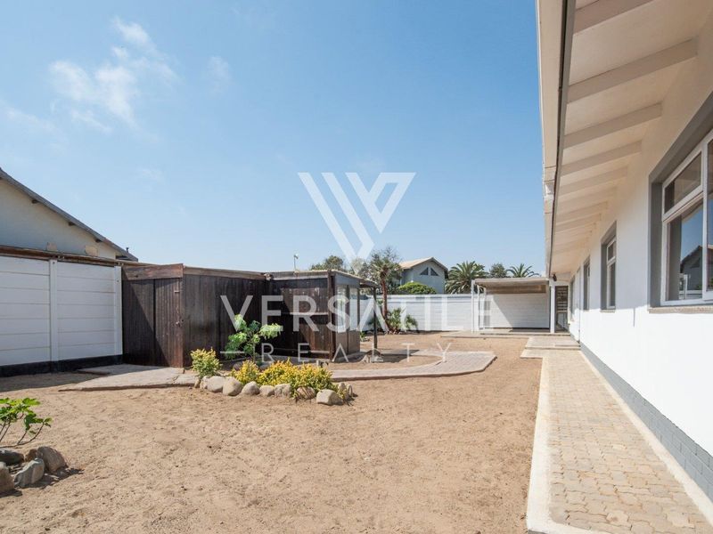 Rear exterior view of the property featuring a paved walkway, sandy yard area, and a timber storage shed against a boundary wall.
