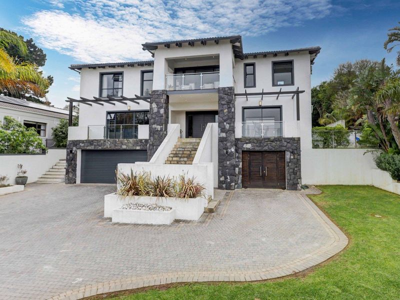 Modern multi-story residence featuring stone cladding, a paved driveway, double garage, and elevated balconies with glass balustrades.