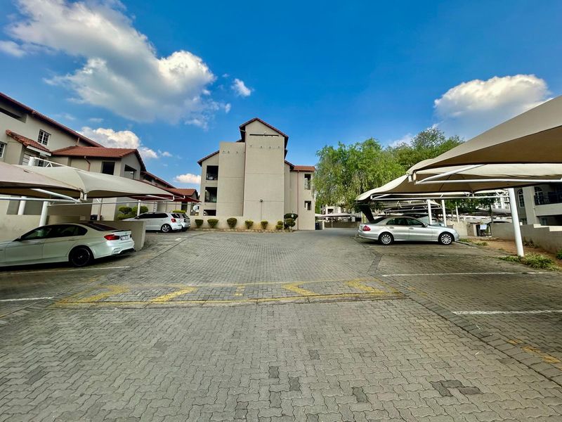 Exterior view of an apartment complex featuring a paved parking area with covered carports and multi-story residential buildings.