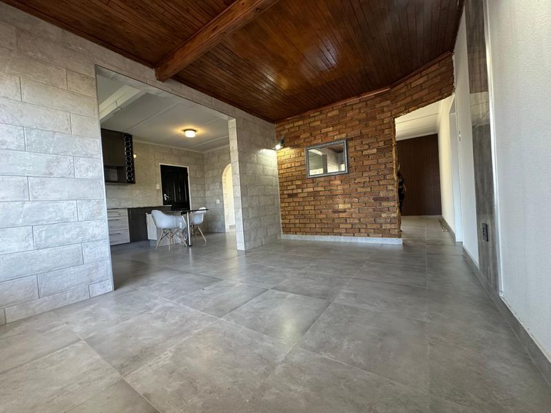 Spacious open-plan living area featuring a warm timber ceiling, exposed brick feature wall, and modern stone-look floor tiles.