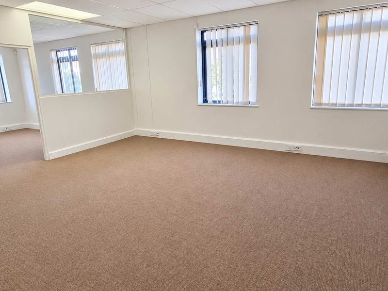 A bright, open-plan office space featuring neutral carpeting, suspended ceilings, and large windows with vertical blinds, offering a clean and professional work environment.