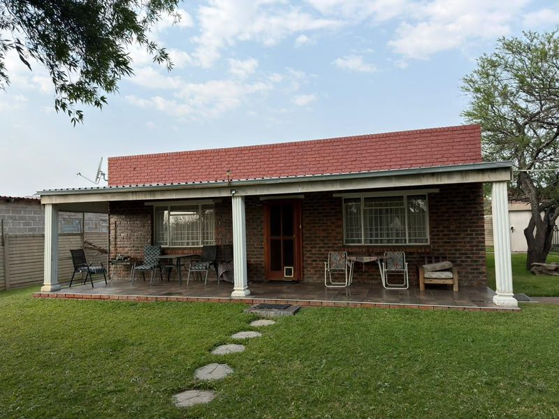 A single-story brick house featuring a covered front patio with tiled flooring, classic columns, and security-barred windows, set within a well-maintained garden.
