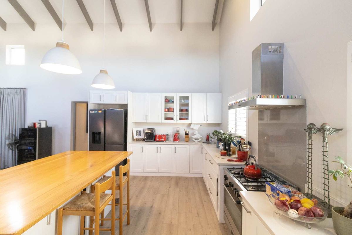 A bright, open-plan kitchen featuring white cabinetry, a wooden breakfast bar, stainless steel gas stove, extractor fan, and high vaulted ceilings.