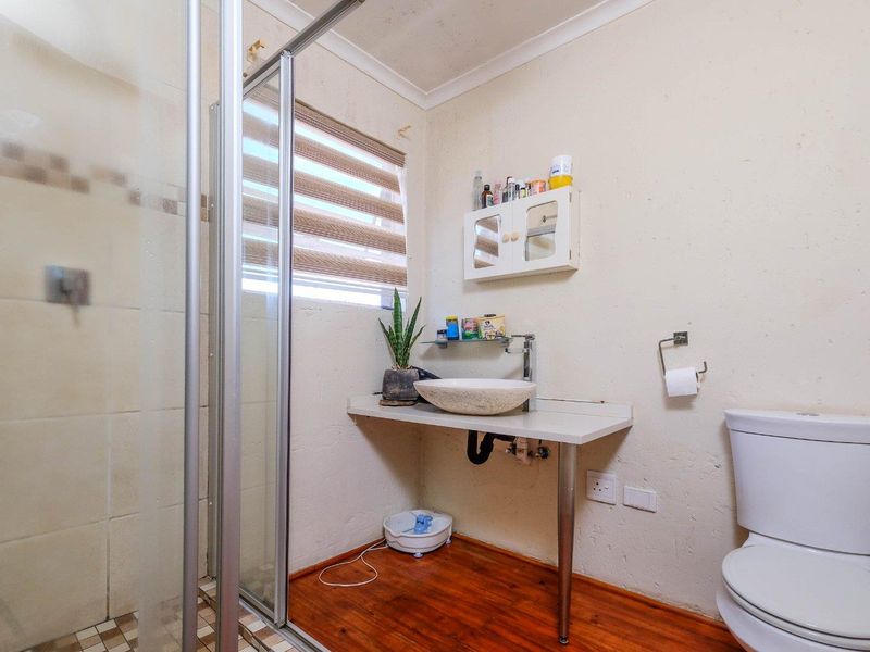 A compact bathroom featuring a shower enclosure, vessel sink on a wall-mounted shelf, and a standard toilet with wooden flooring.
