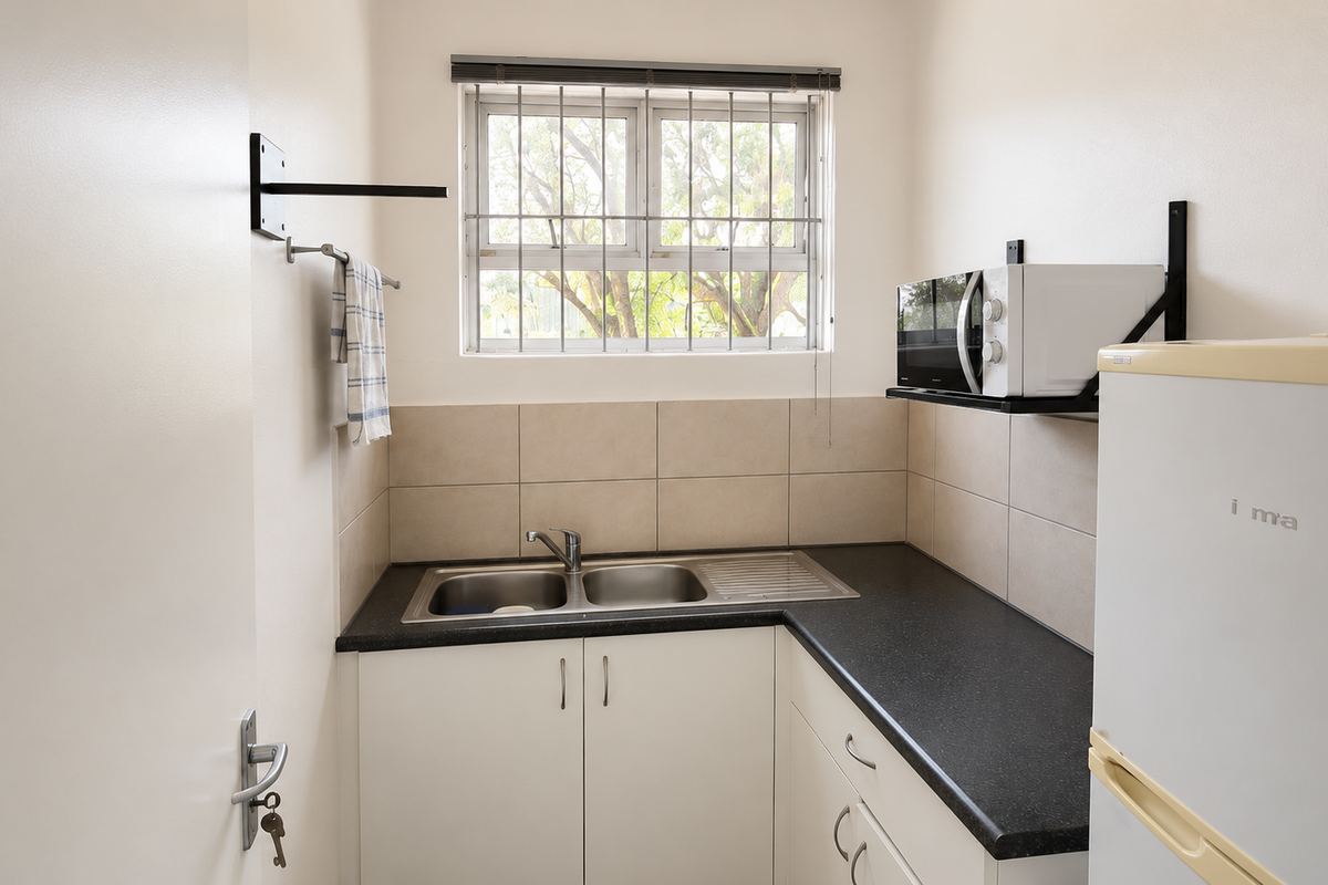 Compact kitchenette featuring a stainless steel double sink, dark laminate countertops, tiled splashback, and built-in cabinetry with security bars on the window.