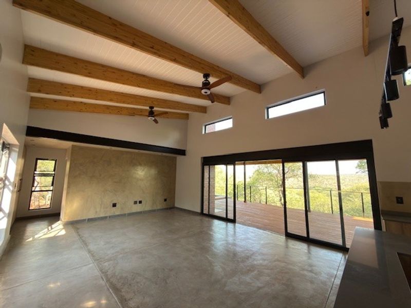 Spacious open-plan living area featuring high ceilings with exposed timber beams, polished concrete floors, and large sliding doors opening to an outdoor deck.