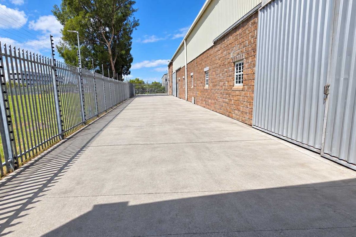 A clean, paved industrial side access area featuring secure steel palisade fencing with electric wiring and durable brick and corrugated metal wall finishes.