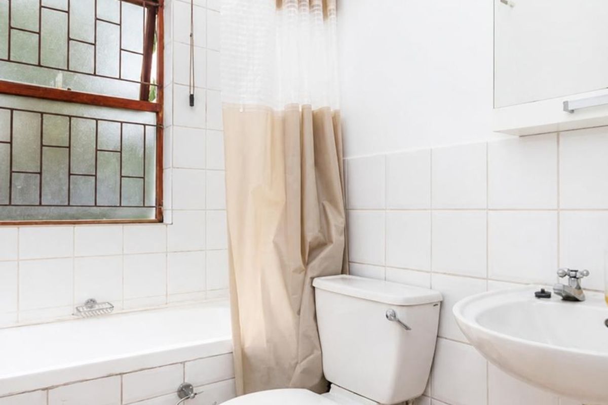 A functional bathroom featuring a bathtub, pedestal sink, and toilet, with white wall tiling and a window fitted with security bars.