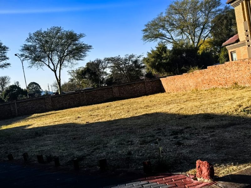 A vacant residential plot featuring a grass lawn, paved driveway area, and a brick perimeter wall.