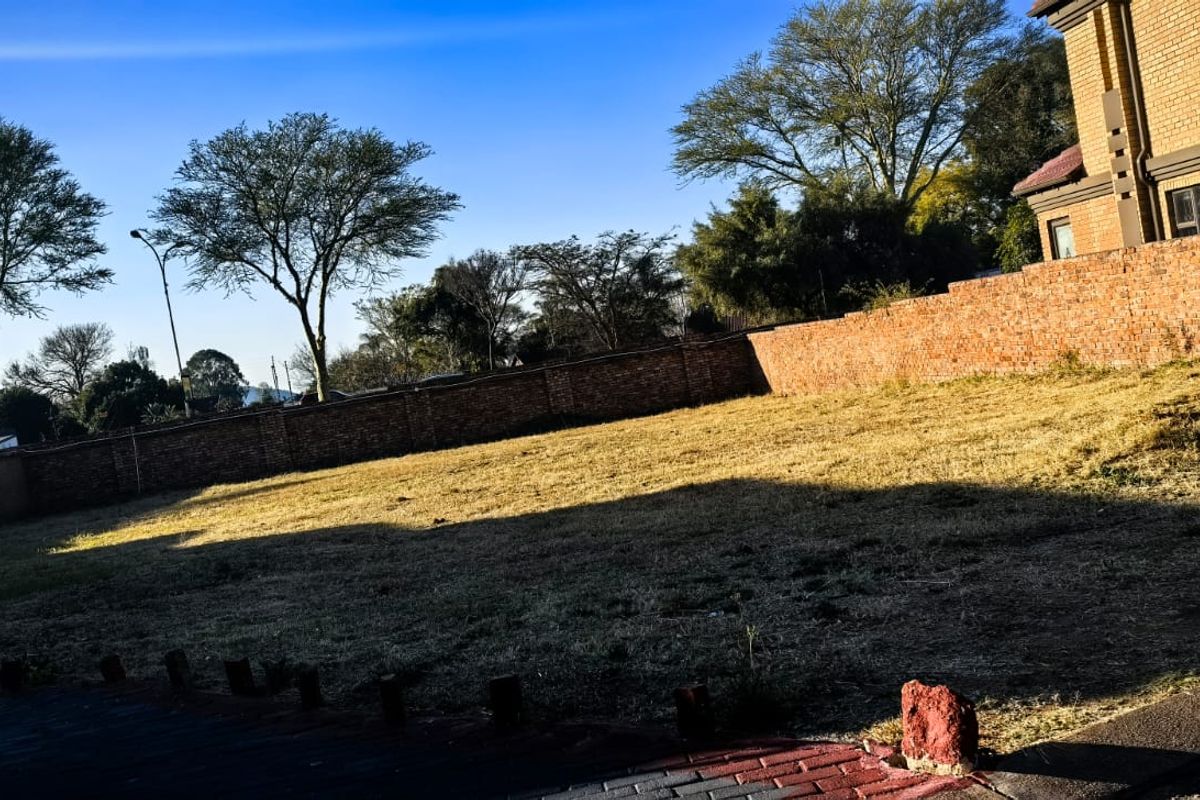 A vacant residential plot featuring a grass lawn, paved driveway area, and a brick perimeter wall.