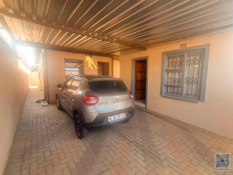 Covered carport area with paved flooring, security window bars, and ventilation brickwork, showing signs of exterior wall wear.