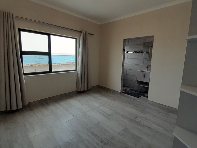 A spacious, empty bedroom featuring modern tiled flooring, large window with natural light, and an adjoining en-suite bathroom with a vanity unit.