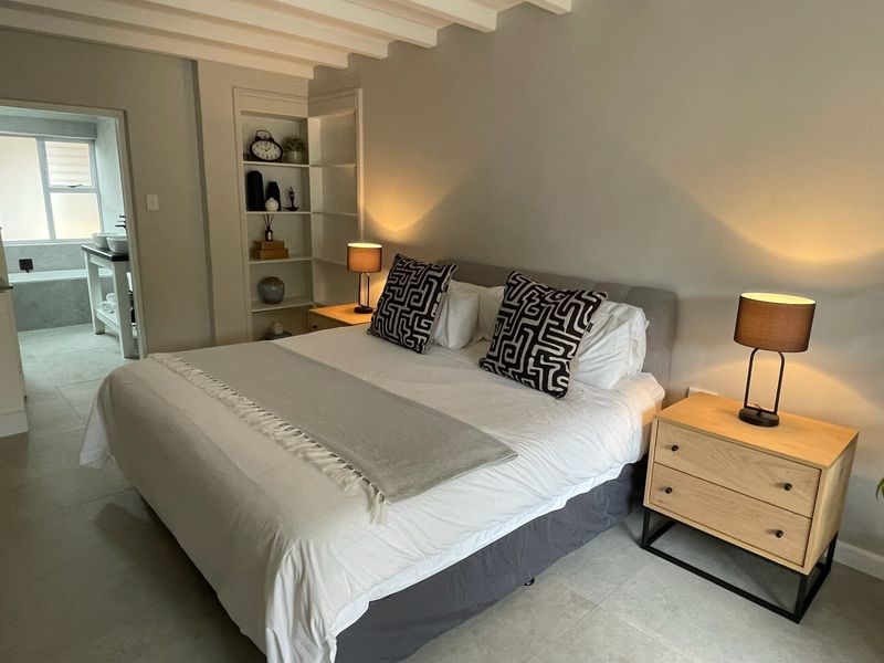 A well-maintained bedroom featuring exposed ceiling beams, built-in shelving, and tiled flooring, with an adjoining bathroom visible in the background.