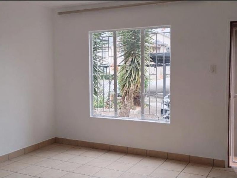A bright, empty living area featuring neutral tiled flooring, security bars on the window, and a secure entrance gate.