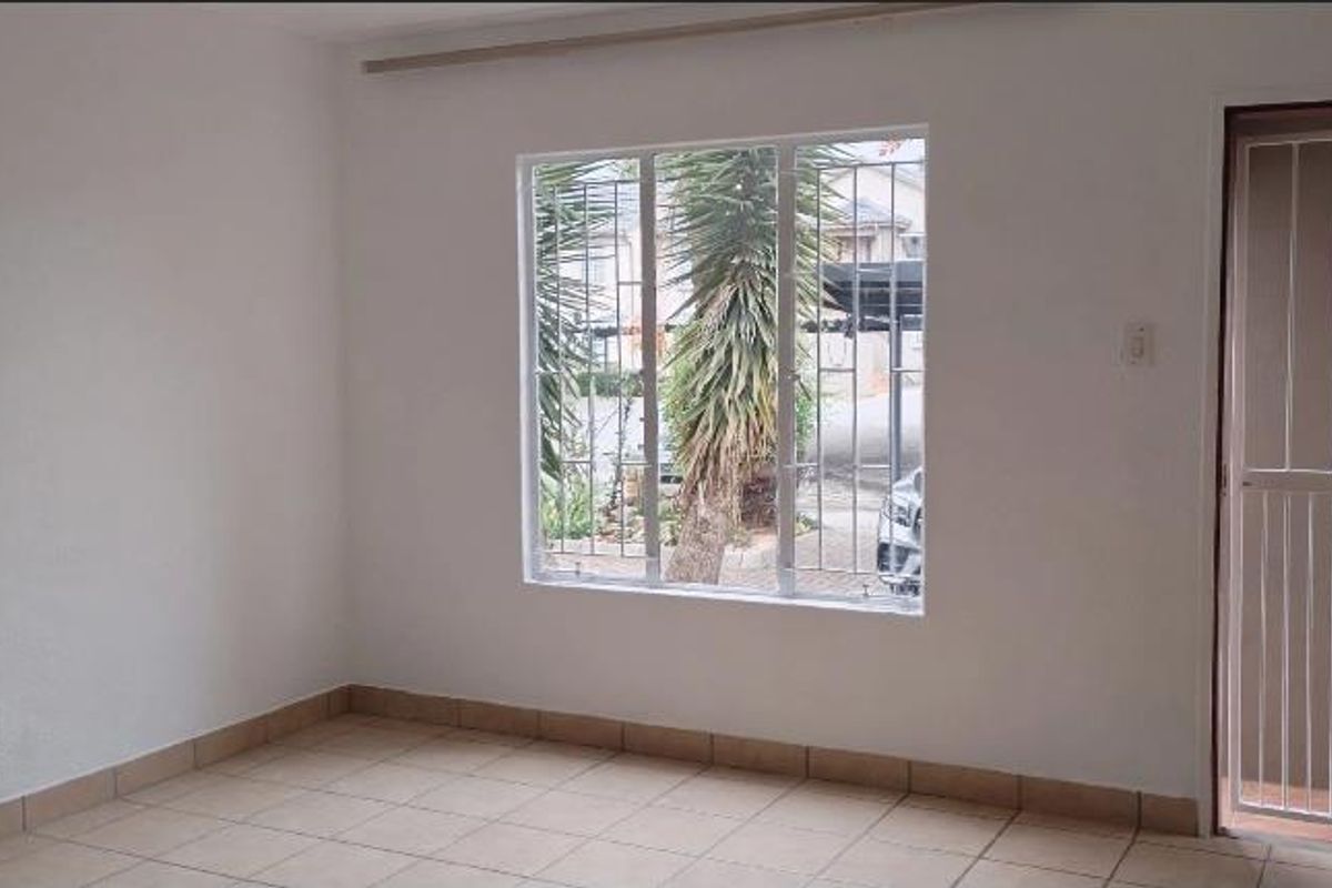 A bright, empty living area featuring neutral tiled flooring, security bars on the window, and a secure entrance gate.
