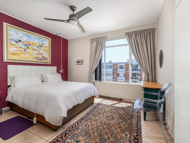 A well-lit bedroom featuring tiled flooring, a ceiling fan, and a large window with views of the surrounding apartment complex.