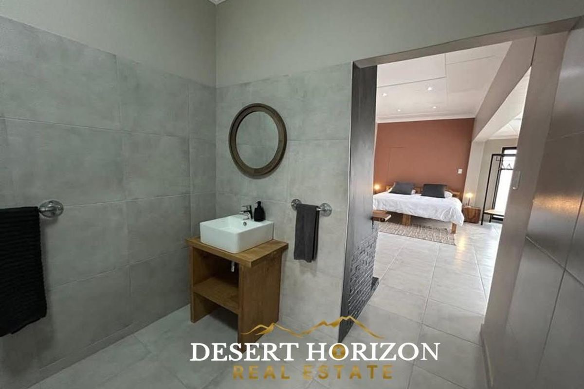 A modern bathroom featuring large-format grey wall tiles, a vessel sink on a wooden vanity, and an open-plan transition into the adjacent bedroom.