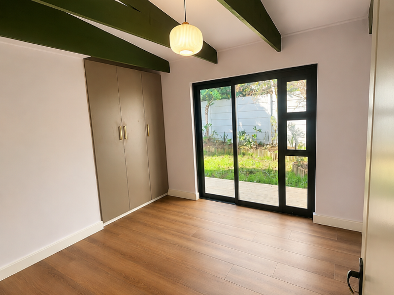 Bright bedroom featuring laminate flooring, built-in cupboards, exposed green timber beams, and a sliding glass door providing direct access to the garden.