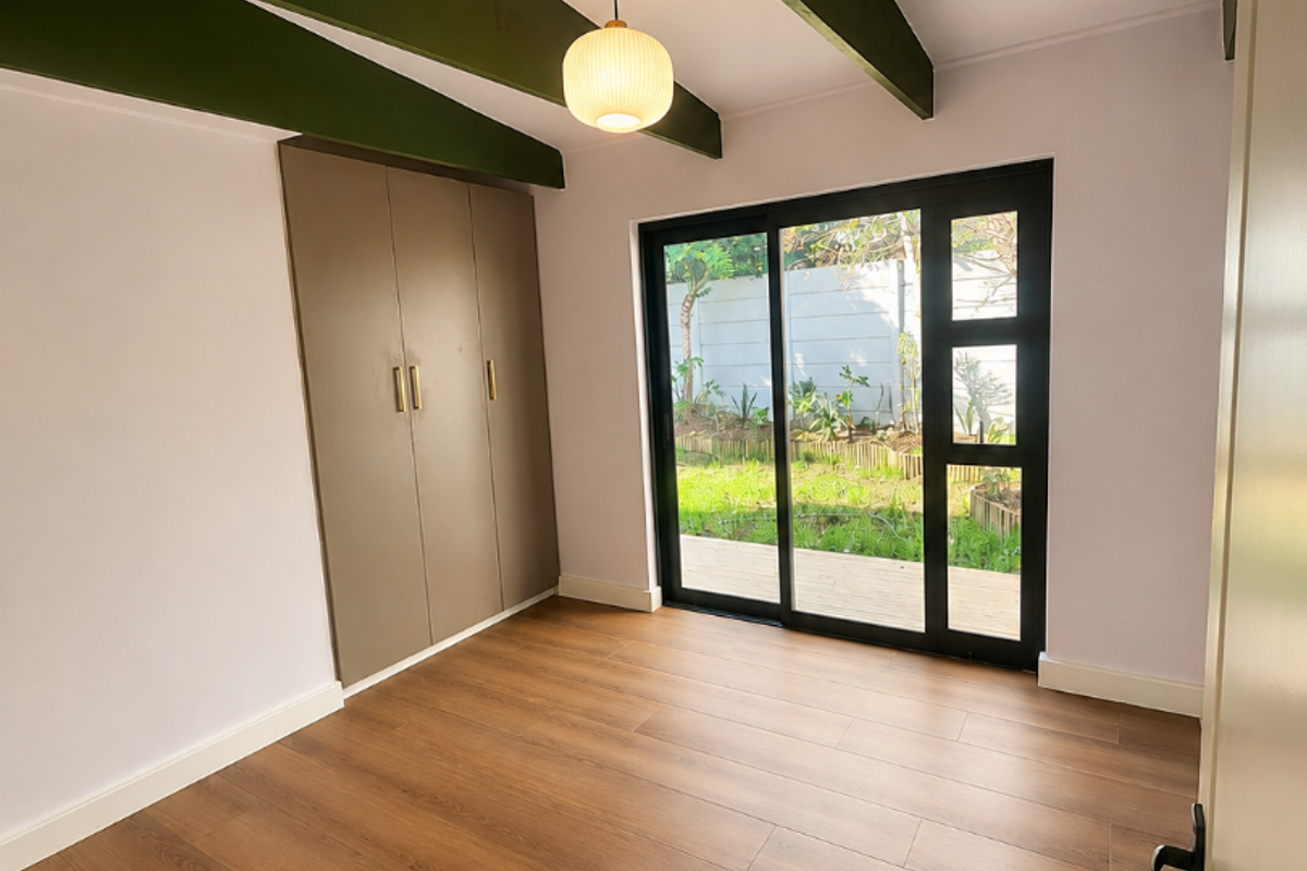 Bright bedroom featuring laminate flooring, built-in cupboards, exposed green timber beams, and a sliding glass door providing direct access to the garden.