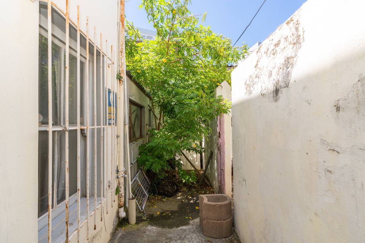 A narrow exterior side passage featuring concrete paving, security bars on windows, and weathered wall finishes requiring maintenance.