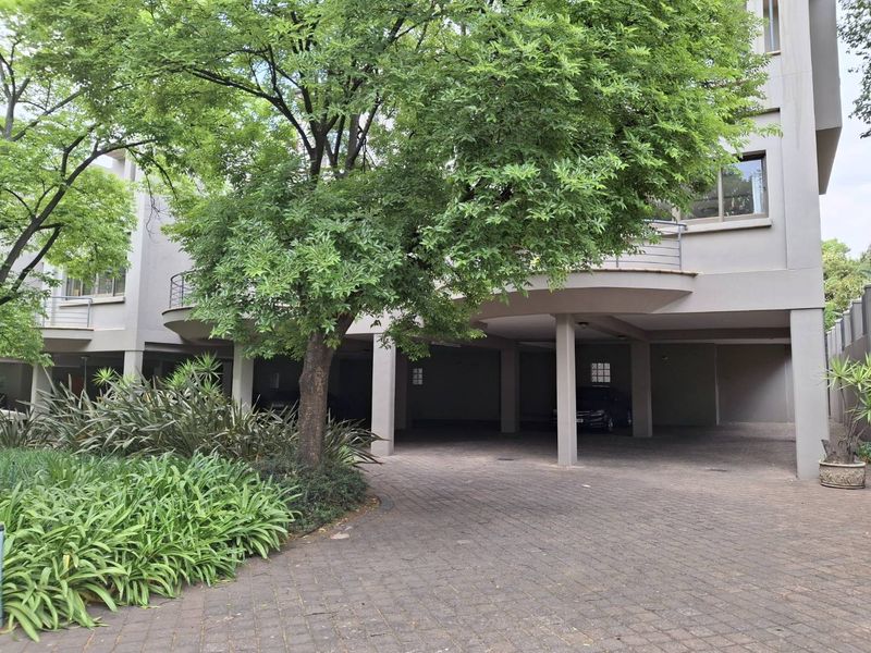 A view of the townhouse exterior featuring a paved driveway, covered parking area, and an upper-level balcony with metal railings.